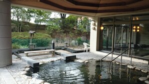 Un área de piscina tranquila al aire libre con bancos de piedra y una pequeña cascada, rodeada por un jardín y un edificio con amplias ventanas.