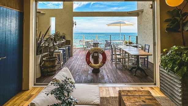An open-plan living area with a view of the sea, featuring a wooden deck, outdoor seating, and potted plants. - L'art de Vie (Matsuyama)