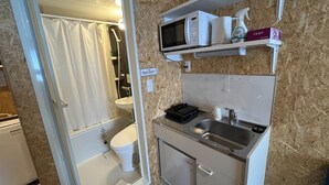A compact bathroom with a shower, toilet, and a small kitchenette with a microwave, sink, and storage shelves.