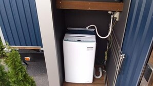 A compact, white laundry machine in a small, enclosed space with a wooden shelf above and a metal rack to the right.