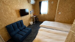 A small room with a blue sofa, two single beds, a desk with a chair, a TV mounted on the wall, and a window with curtains.