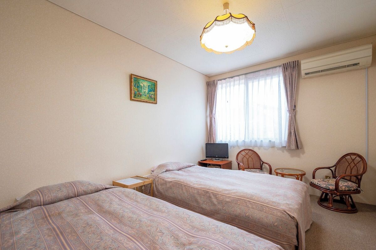 A bedroom with two beds, a desk with a computer, a chair, a lamp, and a painting on the wall.