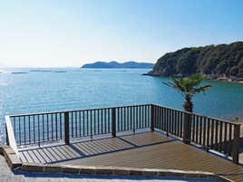 Una terraza de madera con barandilla metálica que da a un mar tranquilo y accidentes geográficos lejanos.