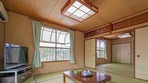 Ein traditioneller japanischer Raum mit Tatami-Böden, einem Fernseher, einem Tisch mit einer Dekoration und verschiebbaren Türen.