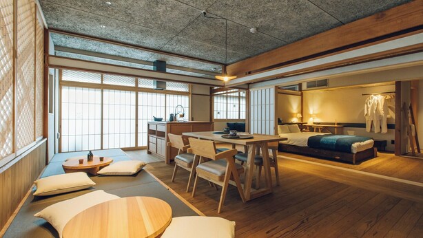 A traditional Japanese-style room with tatami flooring, a low table with cushions, a bed with a blue blanket, and a kitchen area with a sink and counter. - Showcase Hotel Kasane (Gotsu)