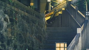A stone staircase with a wooden handrail and a metal railing, leading to a building with a yellow awning and a lit window. - Showcase Hotel Kasane (Gotsu)