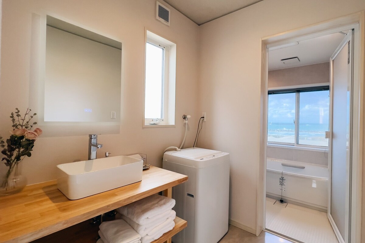 A bathroom with a white sink, a mirror, a window, a washing machine, and a view of the sea.