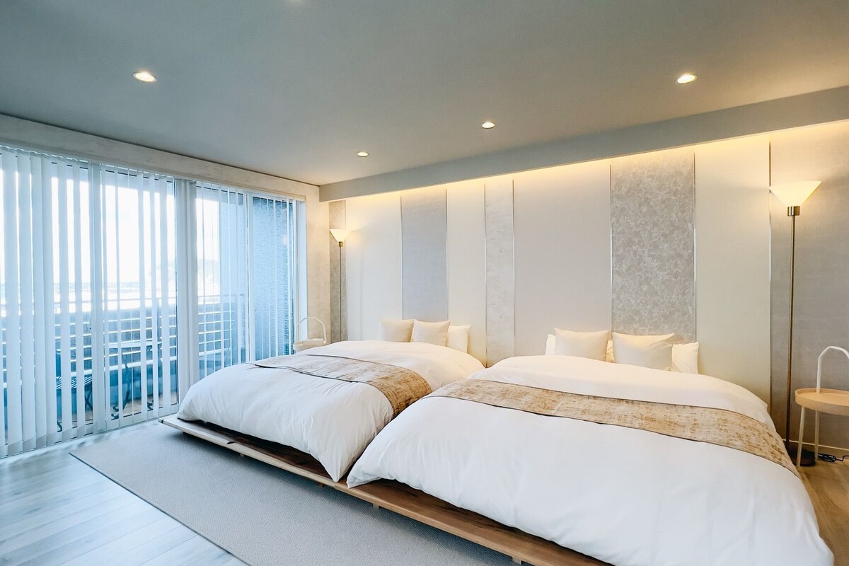 A modern bedroom with two beds, a large window with blinds, and a view of the outside balcony.