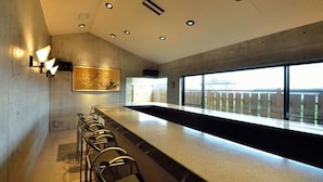 Een moderne bar met een lange toonbank en verschillende barkrukken. Een groot raam biedt uitzicht op het buitengebied.