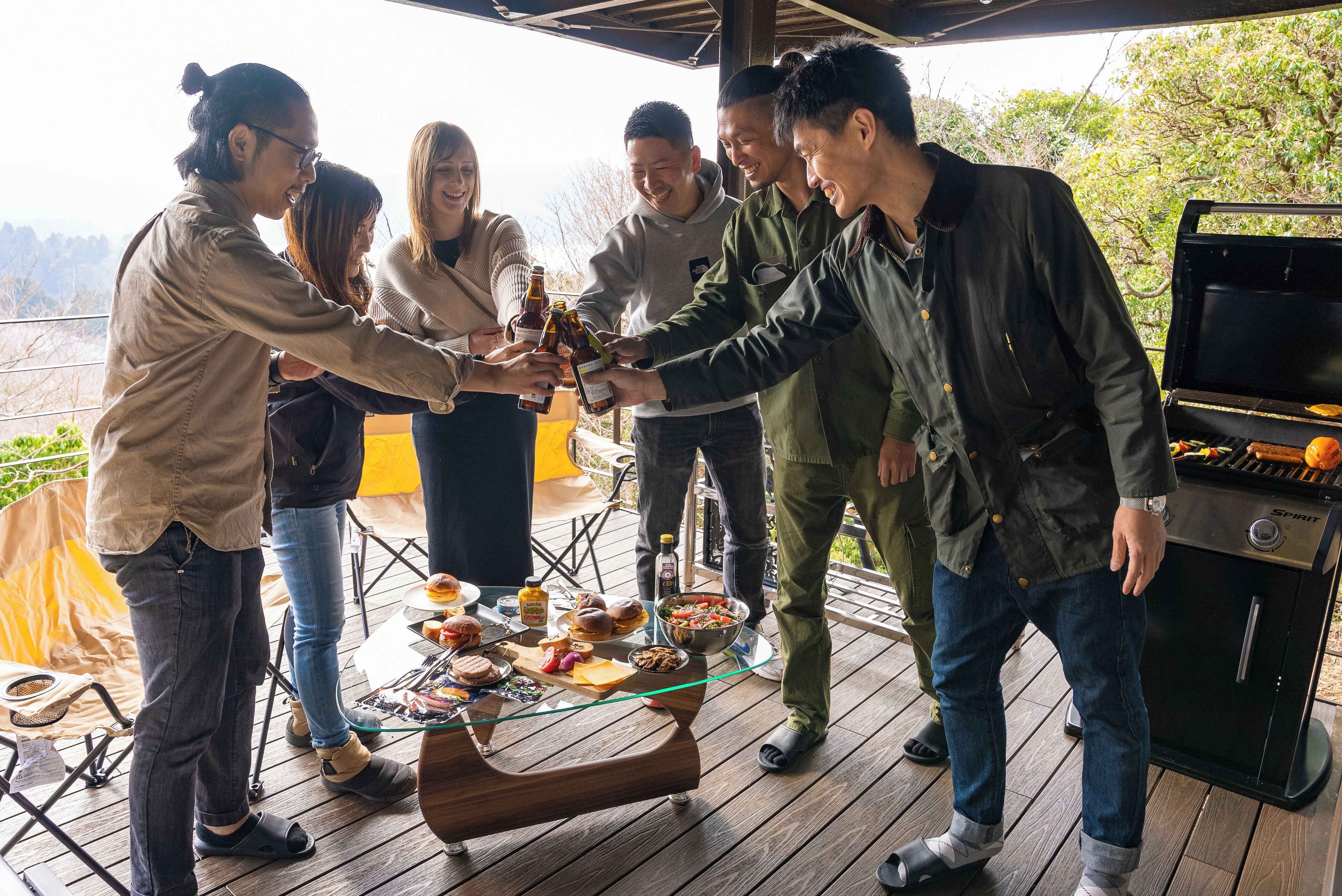 Um grupo de pessoas brindando com garrafas de cerveja em um deck de madeira com churrasqueira e comida.