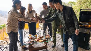 Um grupo de pessoas brindando com garrafas de cerveja em um deck de madeira com churrasqueira e comida.