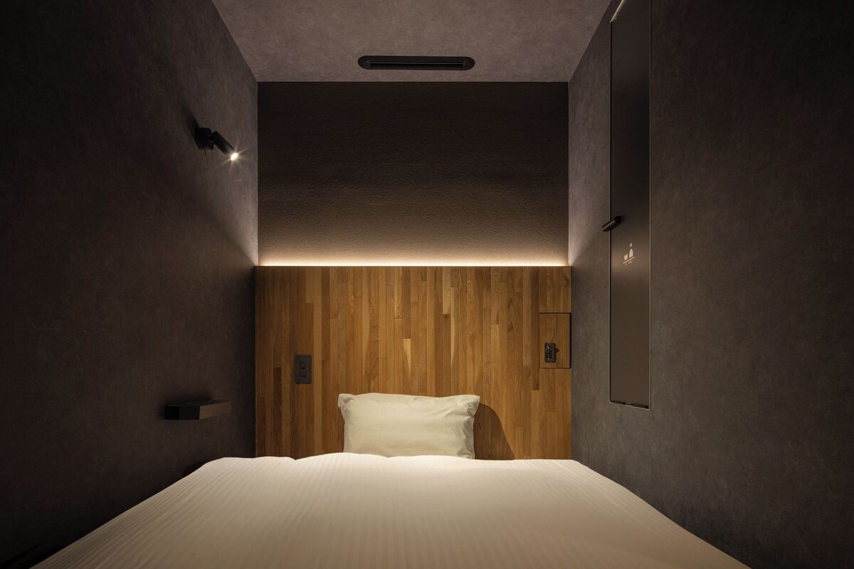 A narrow, dimly lit room with a single bed, wooden panel wall, and a small light fixture above the bed.