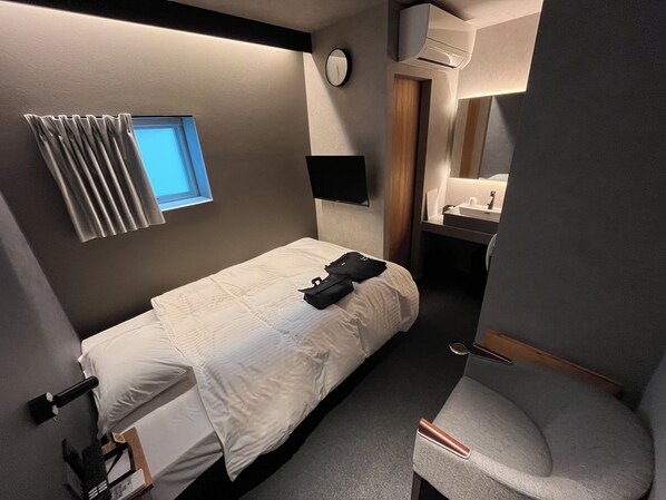 A compact hotel room with a single bed, a small bedside table, a mirror, a window with curtains, a wall-mounted clock, and an air conditioning unit. - Sauna Stay (Setouchi)