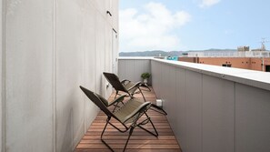A balcony with two lounge chairs, a potted plant, and a view of buildings and hills in the distance. - Sauna Stay (Setouchi)