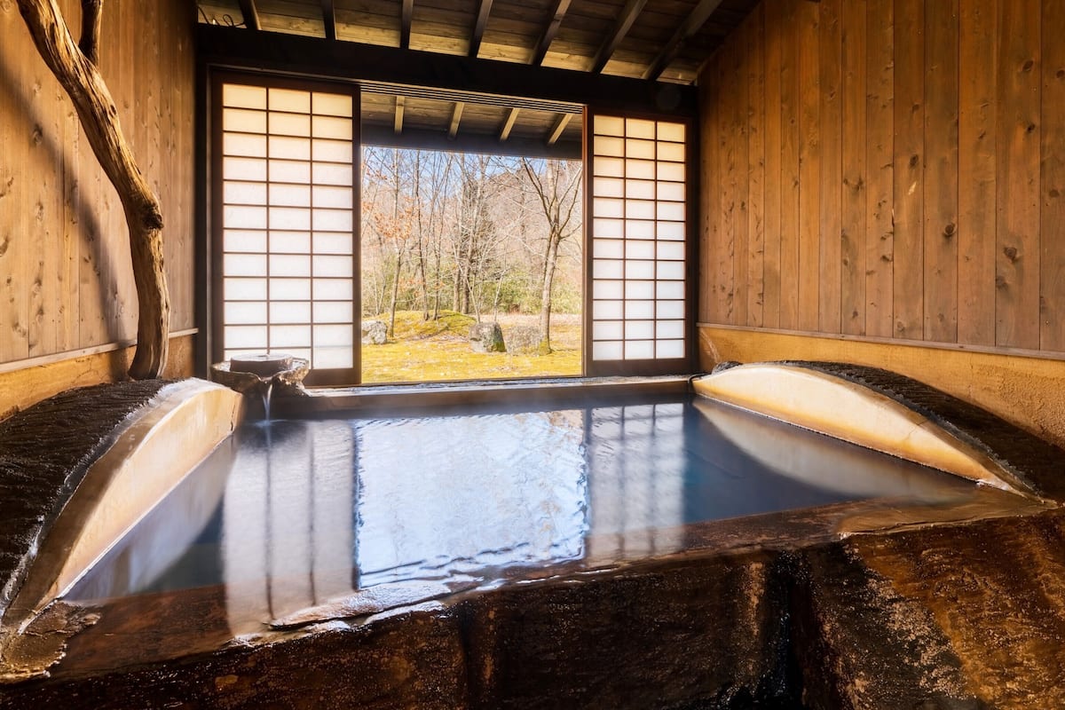 Un onsen japonais traditionnel, avec des murs en bois et une vue sur une forêt, visible à travers des portes coulissantes en papier de riz (shoji).