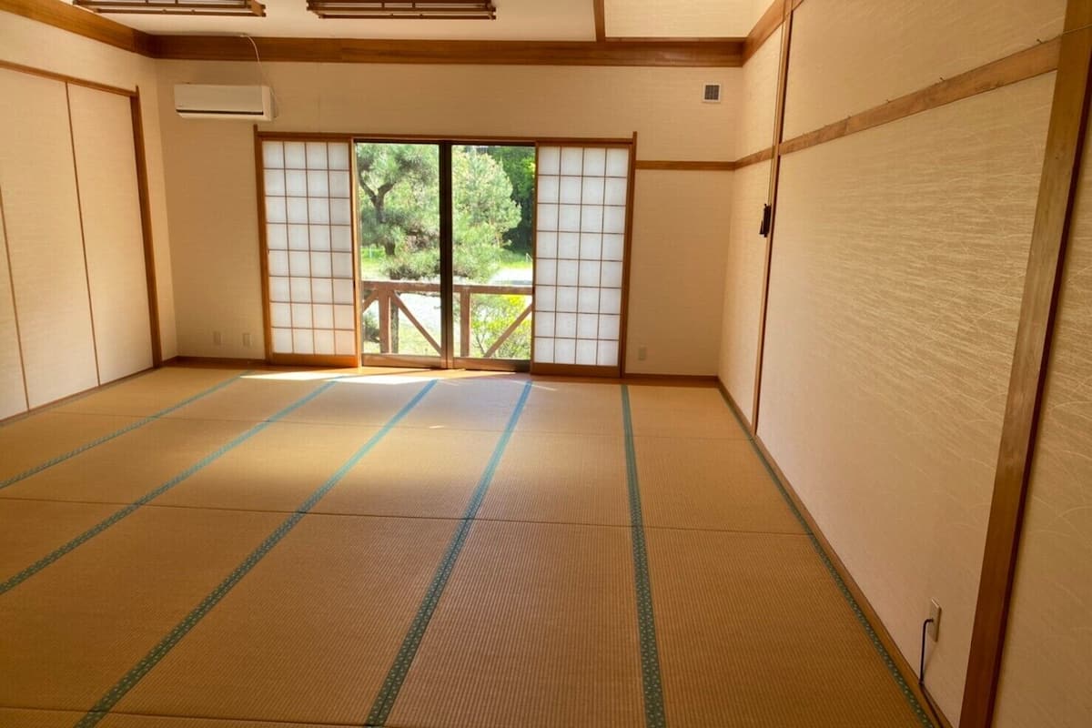 A tatami mat room with sliding doors, a view of greenery outside, and a wooden ceiling.