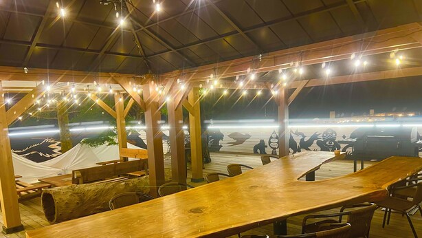 Un espacio de comedor al aire libre con mesas y sillas de madera, luces colgantes y vistas a una parrilla.