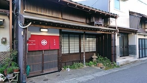 A traditional Japanese building with a red awning, a wooden gate, and a small garden area with plants and a stone lantern. - Hyakunen Machiya Sanotei (Nanao)