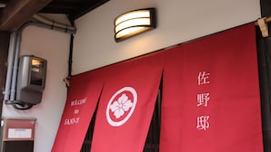 A red banner with white text and a white flower symbol, hanging on a building's exterior. - Hyakunen Machiya Sanotei (Nanao)