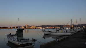 A marina with several boats docked, a calm water body, and a distant cityscape with buildings and hills. - Hyakunen Machiya Sanotei (Nanao)