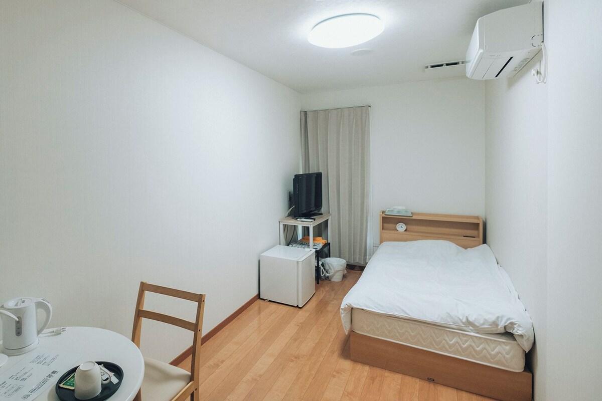 A hotel room with a bed, a desk with a chair, a television, a small table with a kettle, and an air conditioning unit.