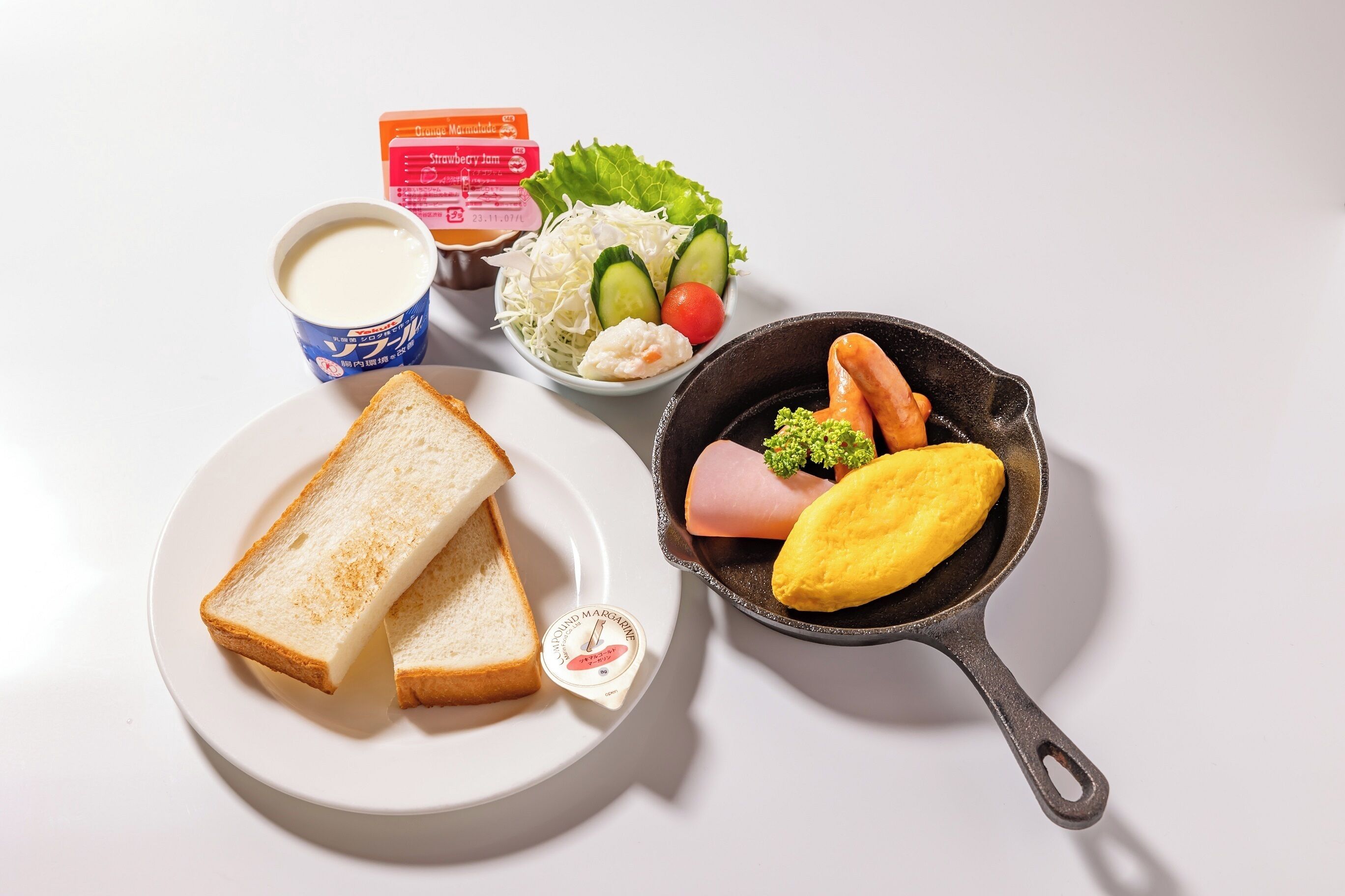 Un plato con dos rebanadas de pan blanco, un recipiente pequeño de mantequilla, un bol de ensalada con lechuga, pepino y tomate, una taza de leche y una sartén de hierro fundido con un trozo de jamón y un huevo.