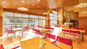 A spacious dining area with wooden tables and red chairs, large windows, and a kitchen in the background. - Riverside Hino (Hino)