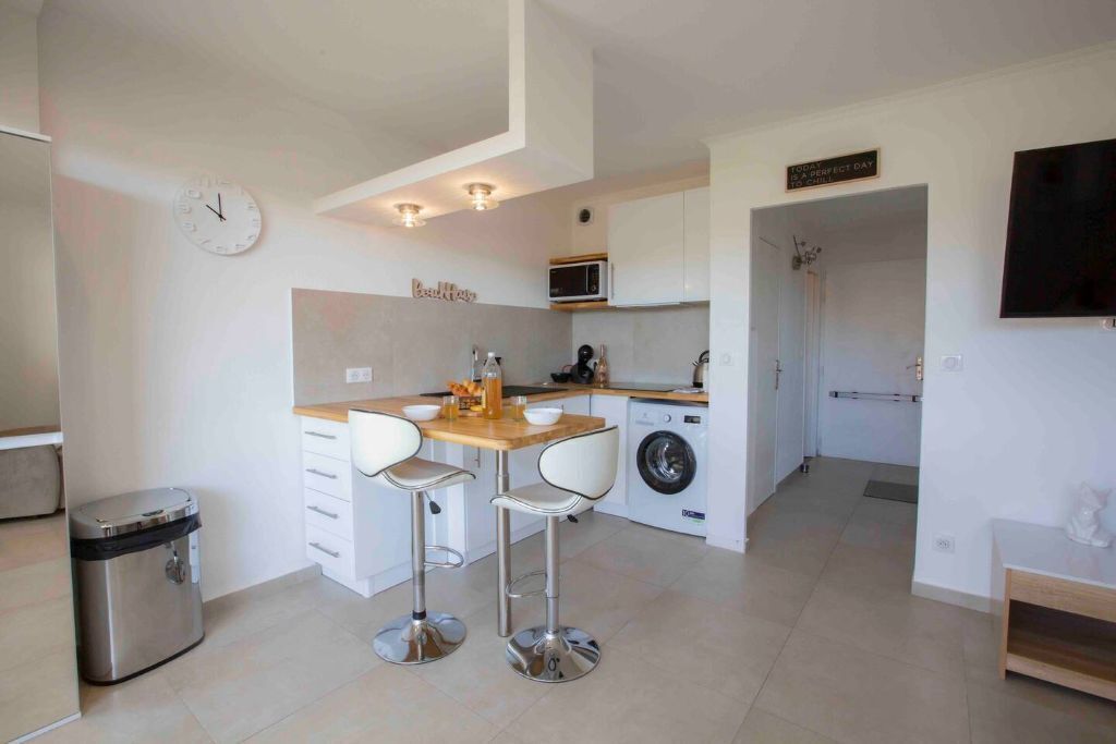 A compact kitchen with a microwave, sink, and washing machine. There are two stools and a small table.