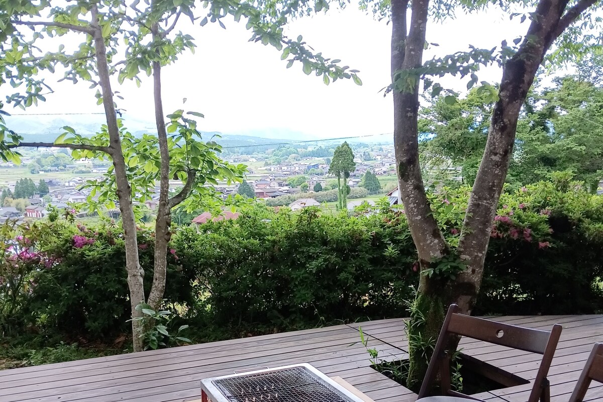 Une terrasse en bois avec une table et des chaises, un barbecue en métal, et une vue sur une ville et des montagnes en arrière-plan.