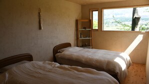 Een slaapkamer met twee bedden, een houten vloer, een raam met uitzicht op groen en een plank met spullen.