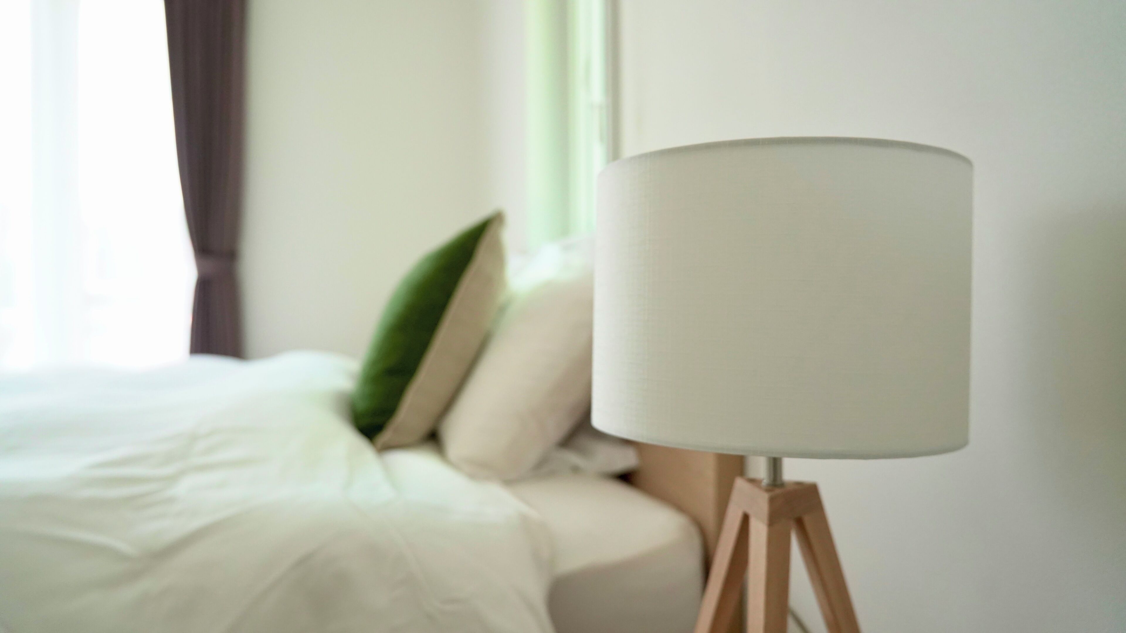 A bed with white bedding, a green pillow, and a white lamp on a wooden stand.