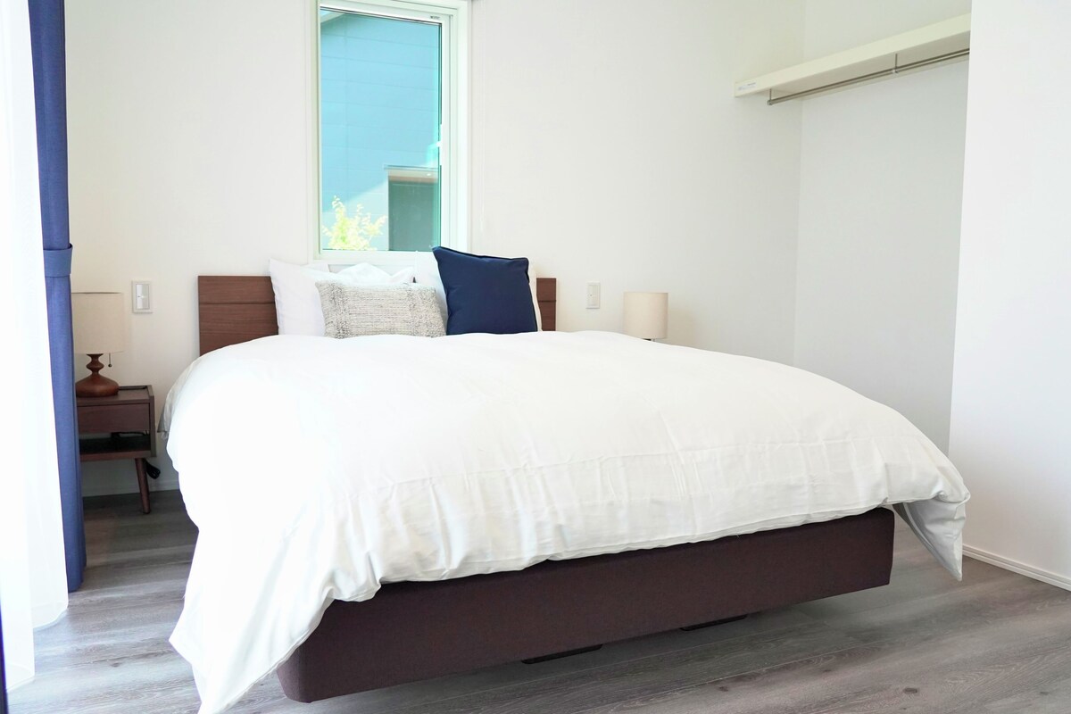 A bedroom with a large bed, a nightstand, a lamp, and a window with a view of greenery.