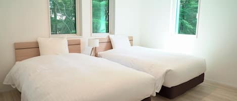 A hotel room with two beds, a wooden headboard, a bedside table with a lamp, and windows with a view of trees.