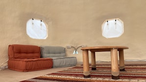 Una habitación acogedora con un rincón de descanso con cojines, una mesa de madera y dos ventanas con cortinas.