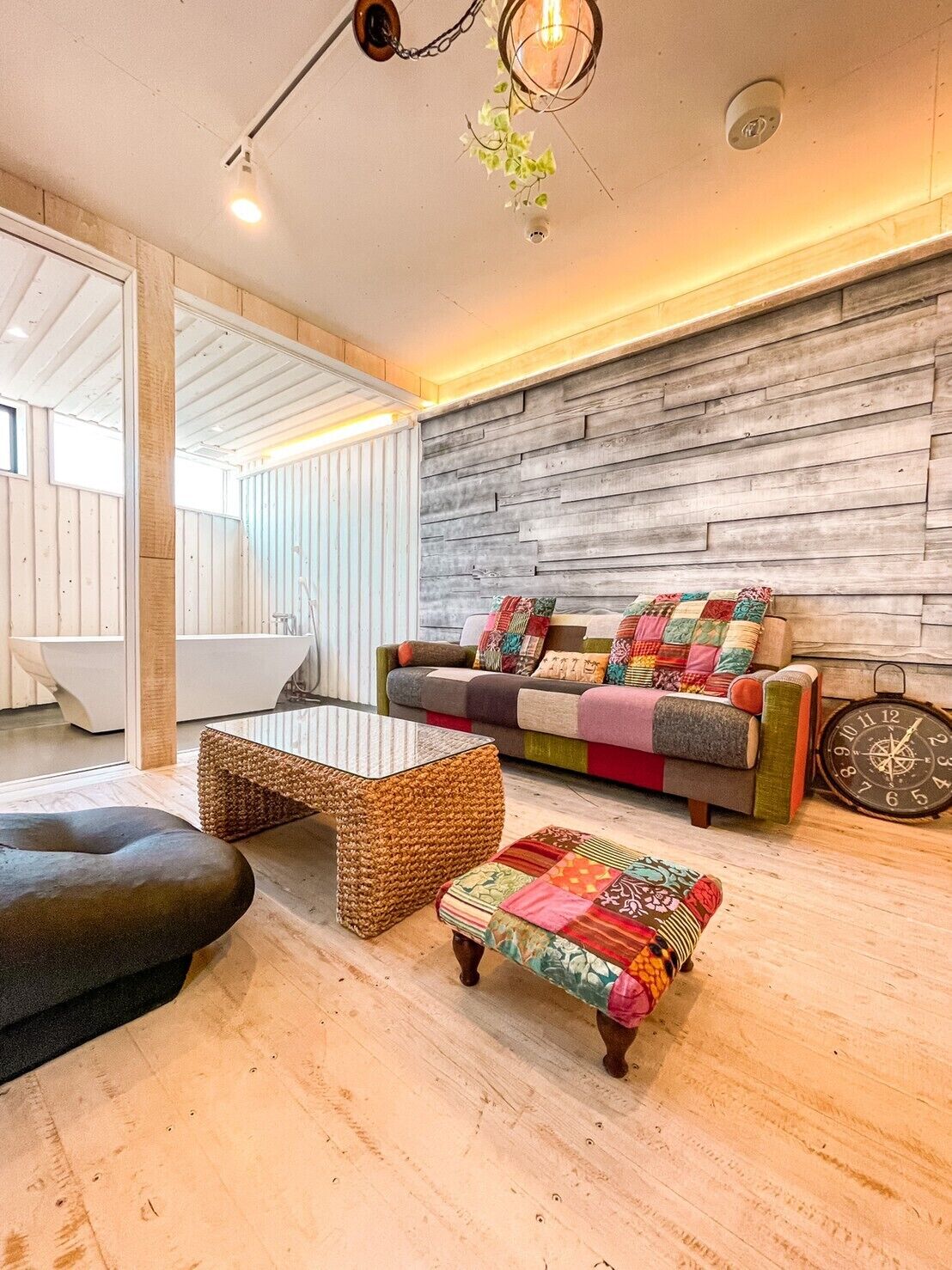 A modern living room with a wooden wall, a colorful sofa, a woven coffee table, and a black ottoman.