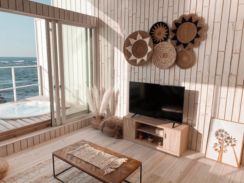 Un amplio salón con una televisión grande, suelo de madera, un mueble de televisión de madera y vistas al océano a través de grandes ventanales.