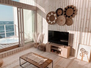 Un vaste espace de vie doté d’un grand téléviseur, de planchers en bois, d’un meuble TV en bois et offrant une vue sur l’océan gr âce à de grandes fenêtres.