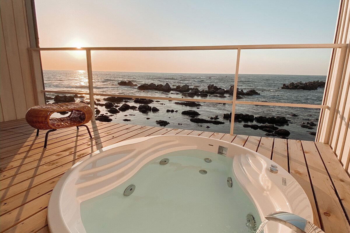 A wooden deck with a hot tub, a wicker chair, and a view of the ocean at sunset.