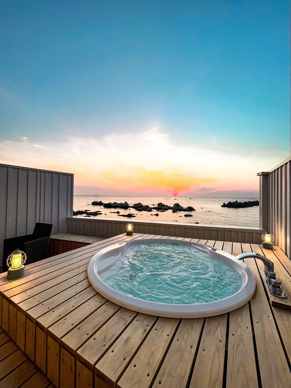 Eine Holzterrasse mit Whirlpool und zwei Laternen mit Blick auf einen Strand mit Felsen und einem Sonnenuntergang.