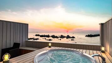 Eine Holzterrasse mit Whirlpool und zwei Laternen mit Blick auf einen Strand mit Felsen und einem Sonnenuntergang.