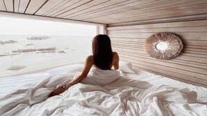 Iemand zit op een bed, met uitzicht op het strand door een raam.