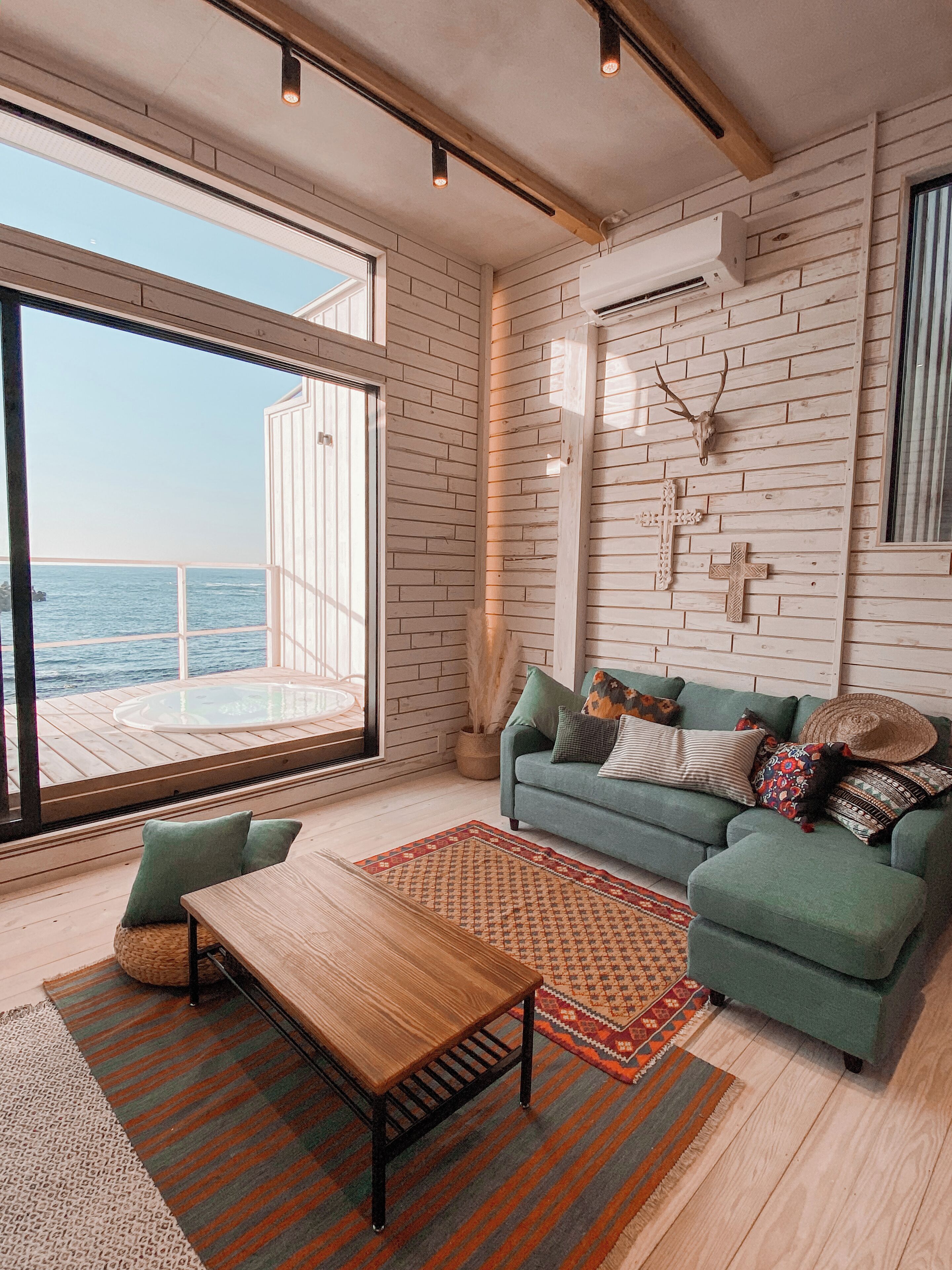 A spacious living area with a large window overlooking the sea, a cozy green sofa, a wooden coffee table, and a rustic wall decor.