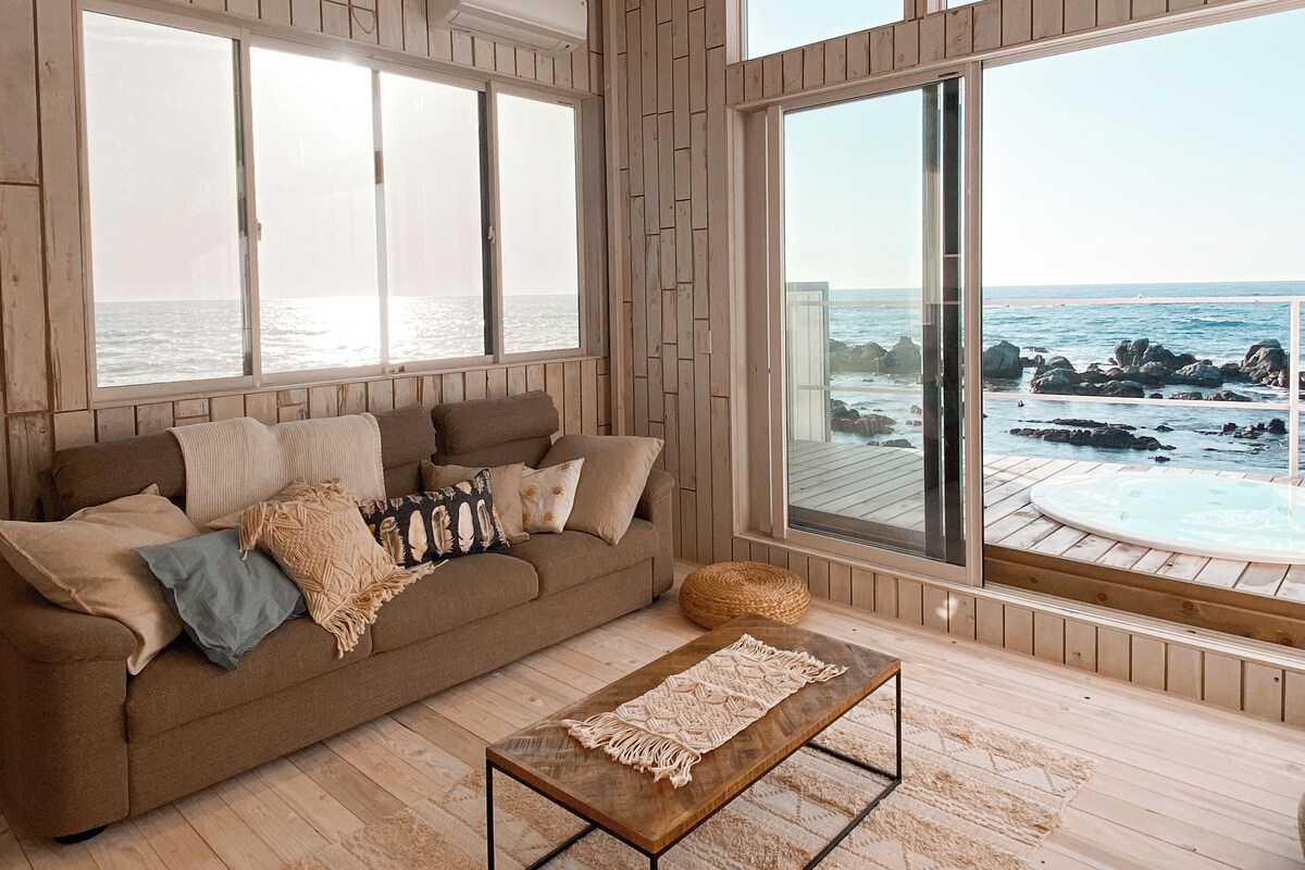 A spacious living room with a large window overlooking the ocean, a cozy brown sofa with cushions, and a wooden coffee table.