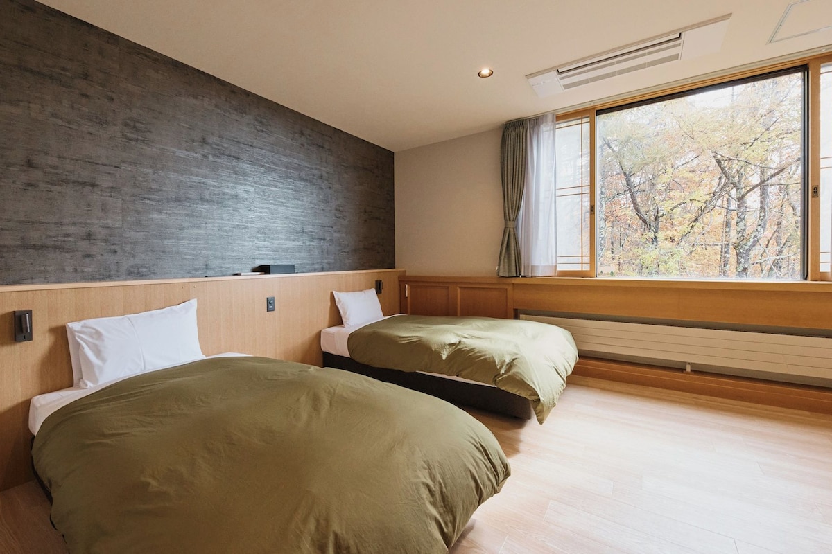 A hotel room with two beds, a large window with curtains, a wooden floor, and a concrete wall.