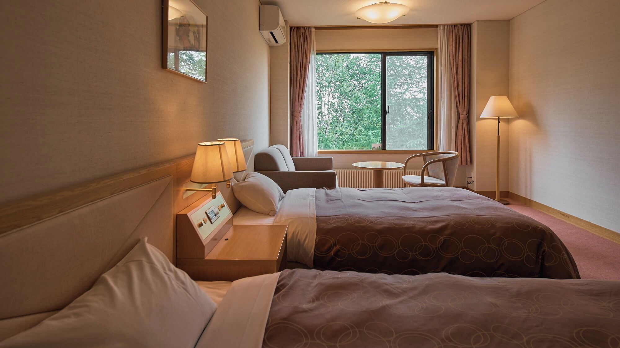 A hotel room with two beds, a small table with a chair, a window with curtains, and a lamp.