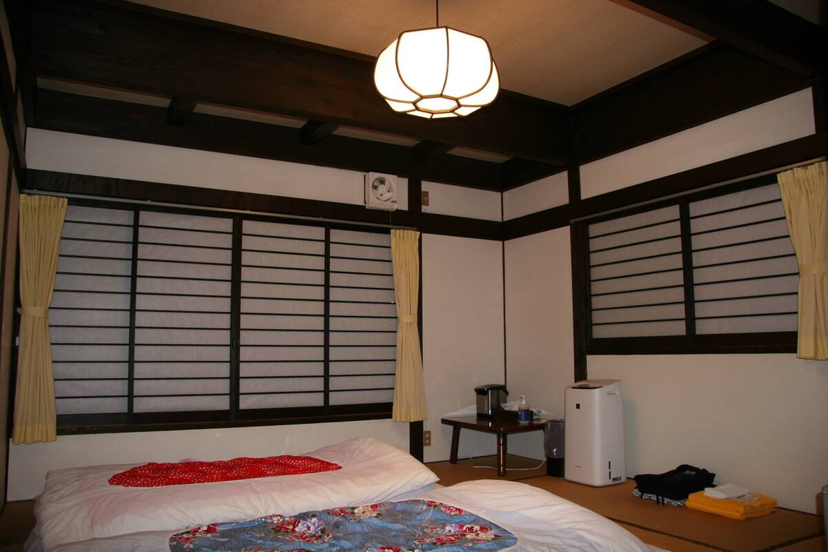 A traditional Japanese-style room with tatami flooring, a bed with a red and blue floral bedspread, a ceiling fan, and a heater.