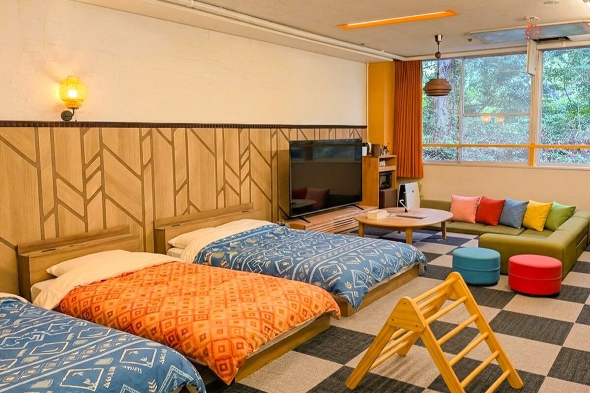 A hotel room with two beds, a sofa, a TV, a window with a view of trees, and a hammock.