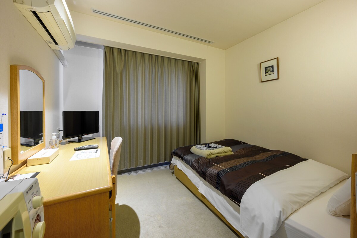 Een hotelkamer met een bed, een bureau met een computer, een stoel, een raam met gordijnen en een foto aan de muur.