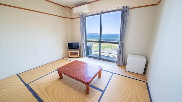 A room with tatami flooring, a wooden table, a television on a stand, a large window with a view of the sea, and an air conditioning unit. - Etoile Oishi (Sumoto)