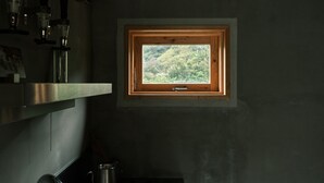 A small window with a wooden frame, a view of a green landscape, and a metal shelf with items. - Negura (Ogimi)